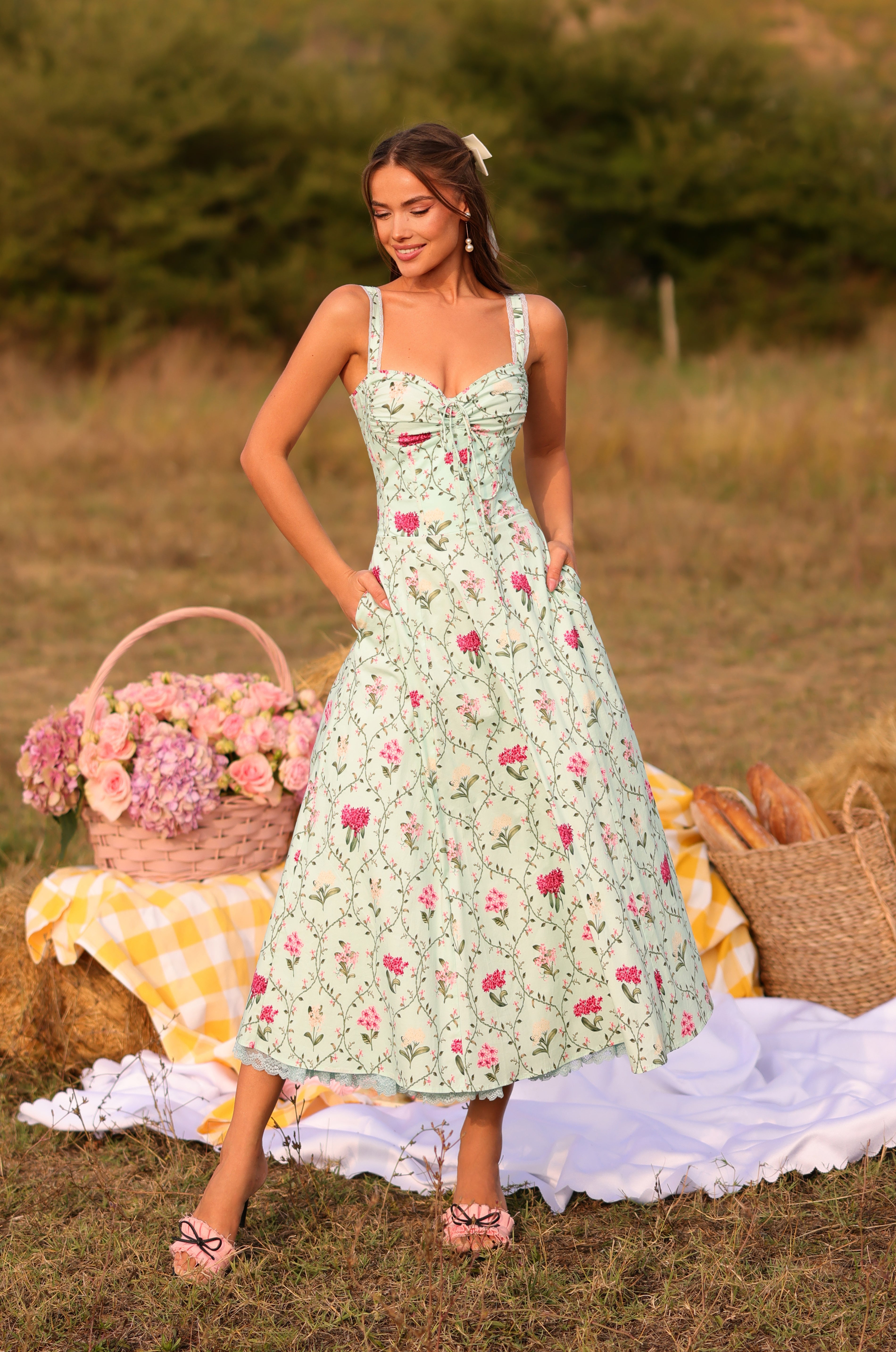 A woman stands outdoors on grass, wearing the Mint Green Floral Casual Midi Dress: Celenia with pockets. In the background, there’s a picnic setup featuring flowers, bread, and a yellow checkered cloth.